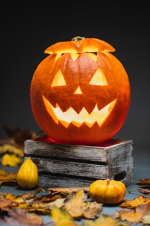 A happy pumpkin on a box with leaves and smaller pumpkins scattered about. The pumpkin is waiting for some fun Halloween Party Games.